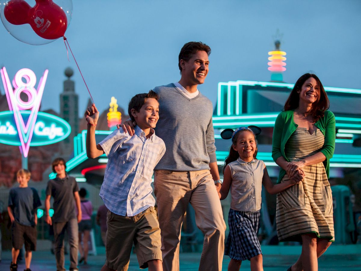 A family of four walks, smiling, with balloons, near a brightly lit neon diner with a 