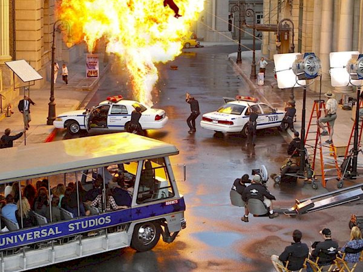 The image shows a movie set with an explosion, police cars, film crew, and a tram labeled 