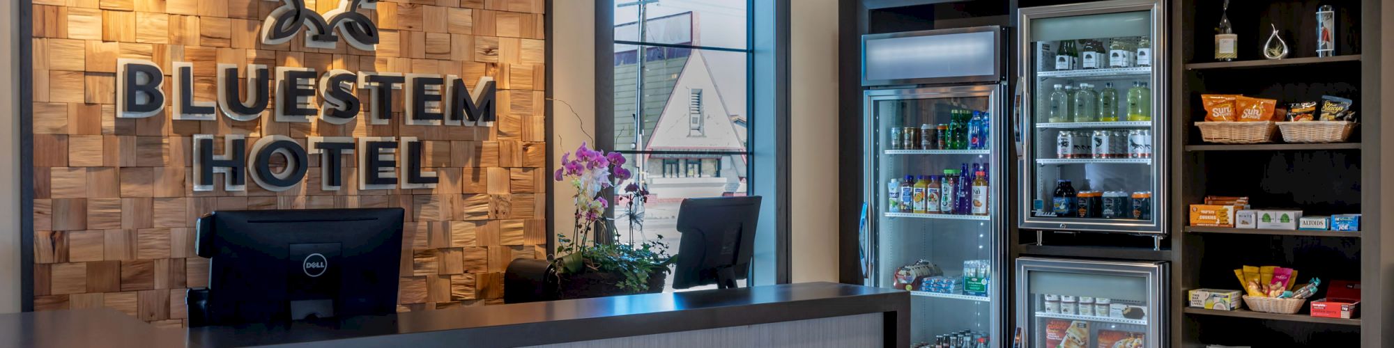 The image shows the reception area of Bluestem Hotel with a front desk, two computer monitors, a floral arrangement, and a well-stocked snack area.