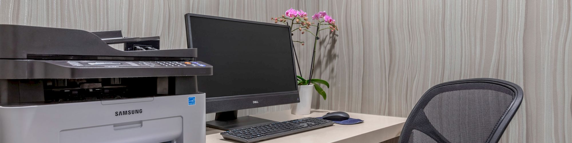 A small office setup with a computer, black chair, Samsung printer, keyboard, and mouse on a white desk, with a potted plant and framed art.