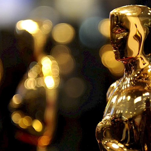 The image shows close-up views of golden statuettes, commonly known as Oscars, against a blurred background with shimmering lights, creating a bokeh effect.