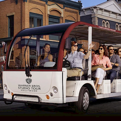 A group of people is seated on a Warner Bros. Studio Tour tram, wearing headphones and enjoying the ride through a film studio set area in Hollywood.