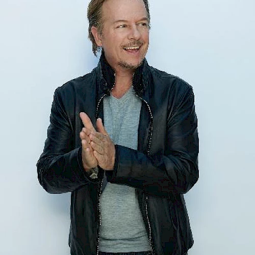 A person is wearing a black jacket and gray shirt, standing against a light background and smiling with hands clasped.