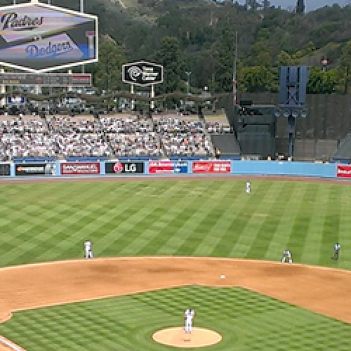 A baseball game is in progress at a stadium with a large crowd, green field, and scoreboards displaying advertisements and game information.