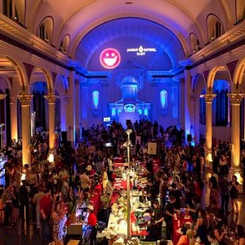 A large crowd gathers inside a grand hall with high ceilings and ornate columns, illuminated by blue and yellow lighting, creating a festive atmosphere.