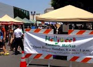 People are walking through a farmers' market with various colorful canopies. A banner indicates it's the Hollywood Farmers' Market, open Sundays.