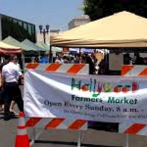 People are walking through a farmers' market with various colorful canopies. A banner indicates it's the Hollywood Farmers' Market, open Sundays.