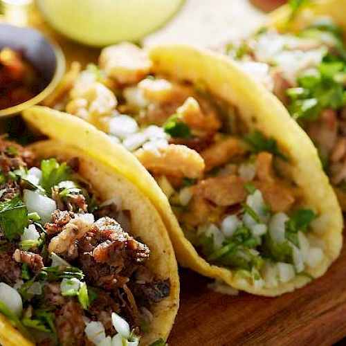 Three tacos filled with different meats, garnished with chopped onions and cilantro, and served with a side of salsa on a wooden board.