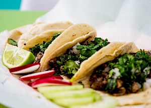 The image shows four tacos filled with meat, chopped onions, and cilantro, garnished with lime wedges, sliced radishes, and cucumber slices.