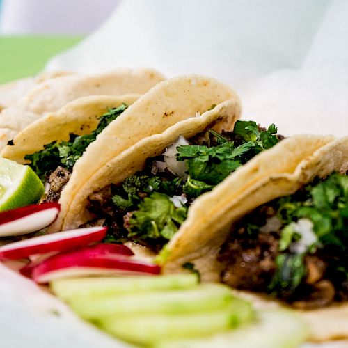 The image shows four tacos filled with meat, chopped onions, and cilantro, garnished with lime wedges, sliced radishes, and cucumber slices.