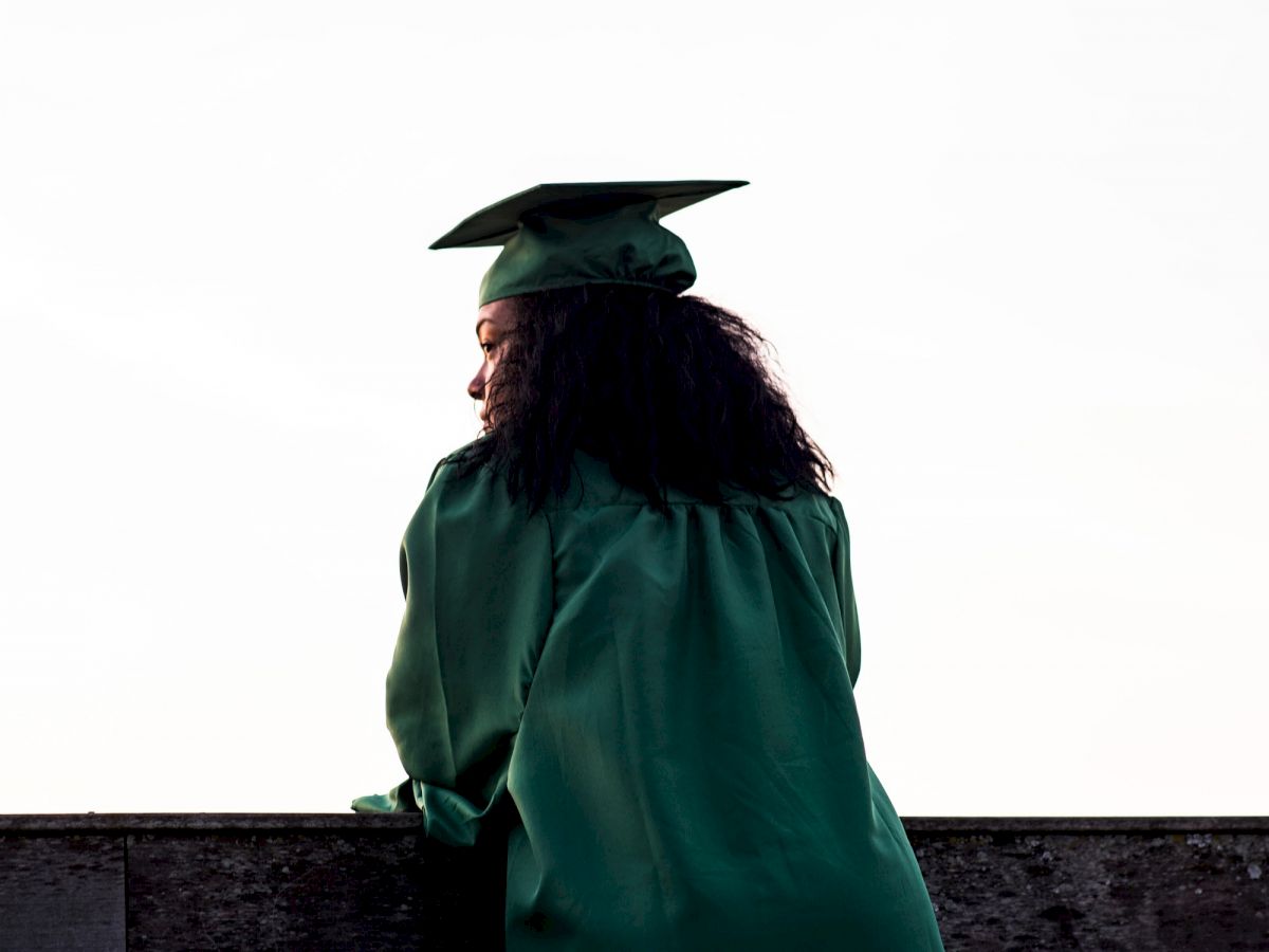 A person in a green graduation gown and cap is standing against a ledge, facing away from the camera.