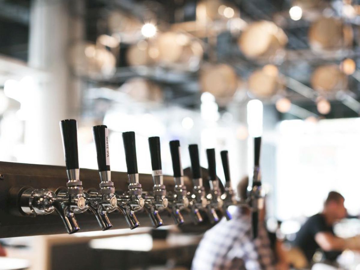 The image shows a row of beer taps in a modern bar setting with blurred background, featuring patrons and overhead lighting, creating a casual atmosphere.