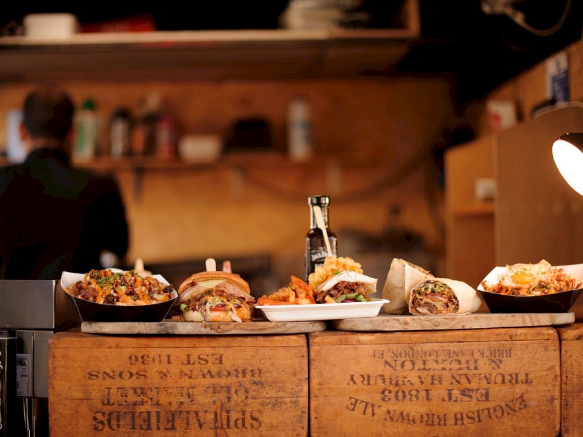 The image shows various types of fast food, like burgers and wraps, displayed on wooden crates, with a dimly lit kitchen visible in the background.