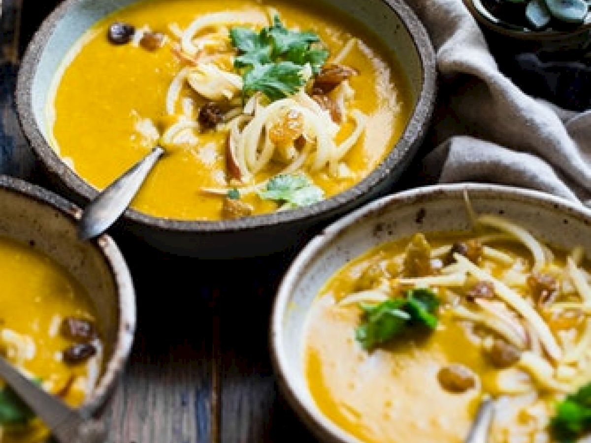 The image shows bowls of creamy soup garnished with herbs, wooden table setting, bread pieces, salt and pepper shakers, and succulent plants.