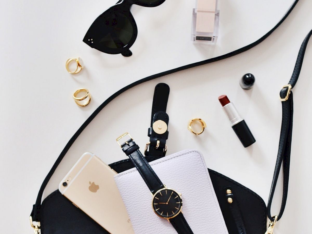 The image shows a black quilted purse with sunglasses, foundation, lipstick, rings, a smartphone, watch, and a notebook.