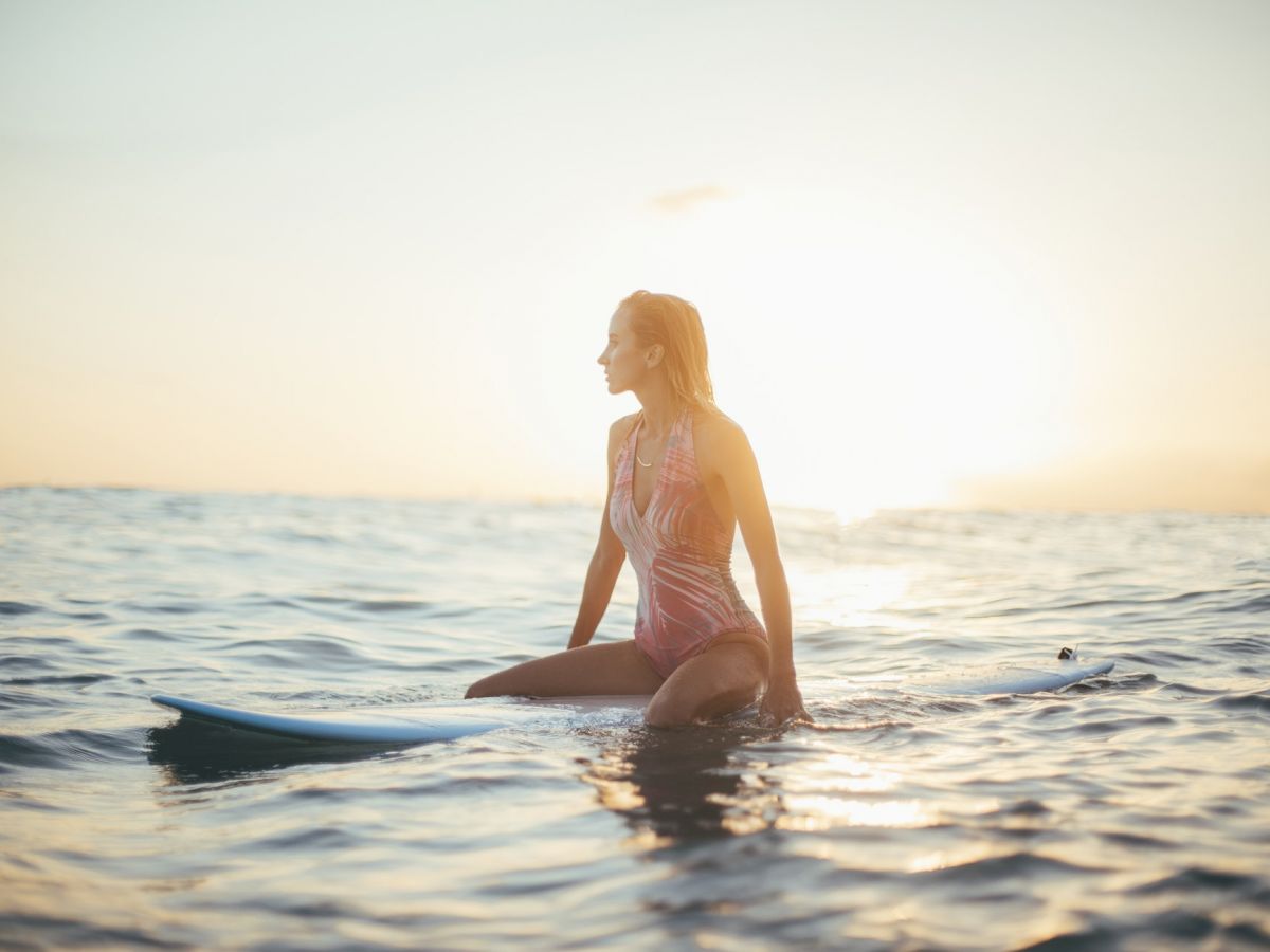 A person wearing a swimsuit sits on a surfboard in the ocean during a sunset, with the sun casting a warm glow over the scene.