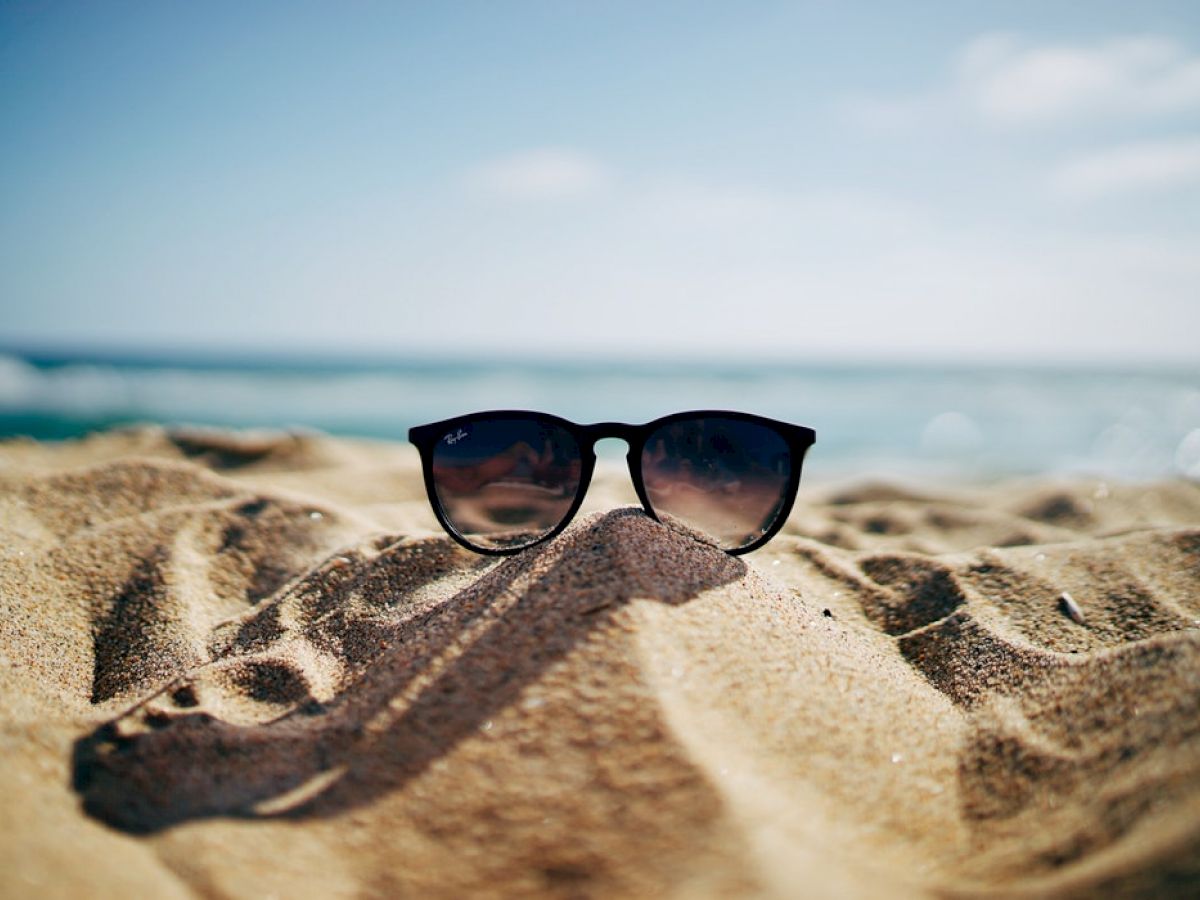 A pair of sunglasses is placed on sand at a beach with the ocean and sky in the background. The scene suggests a relaxed, sunny day.