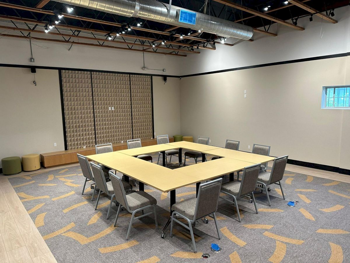 This image shows a conference room with a square arrangement of tables and chairs, a patterned carpet, and exposed ceiling beams.