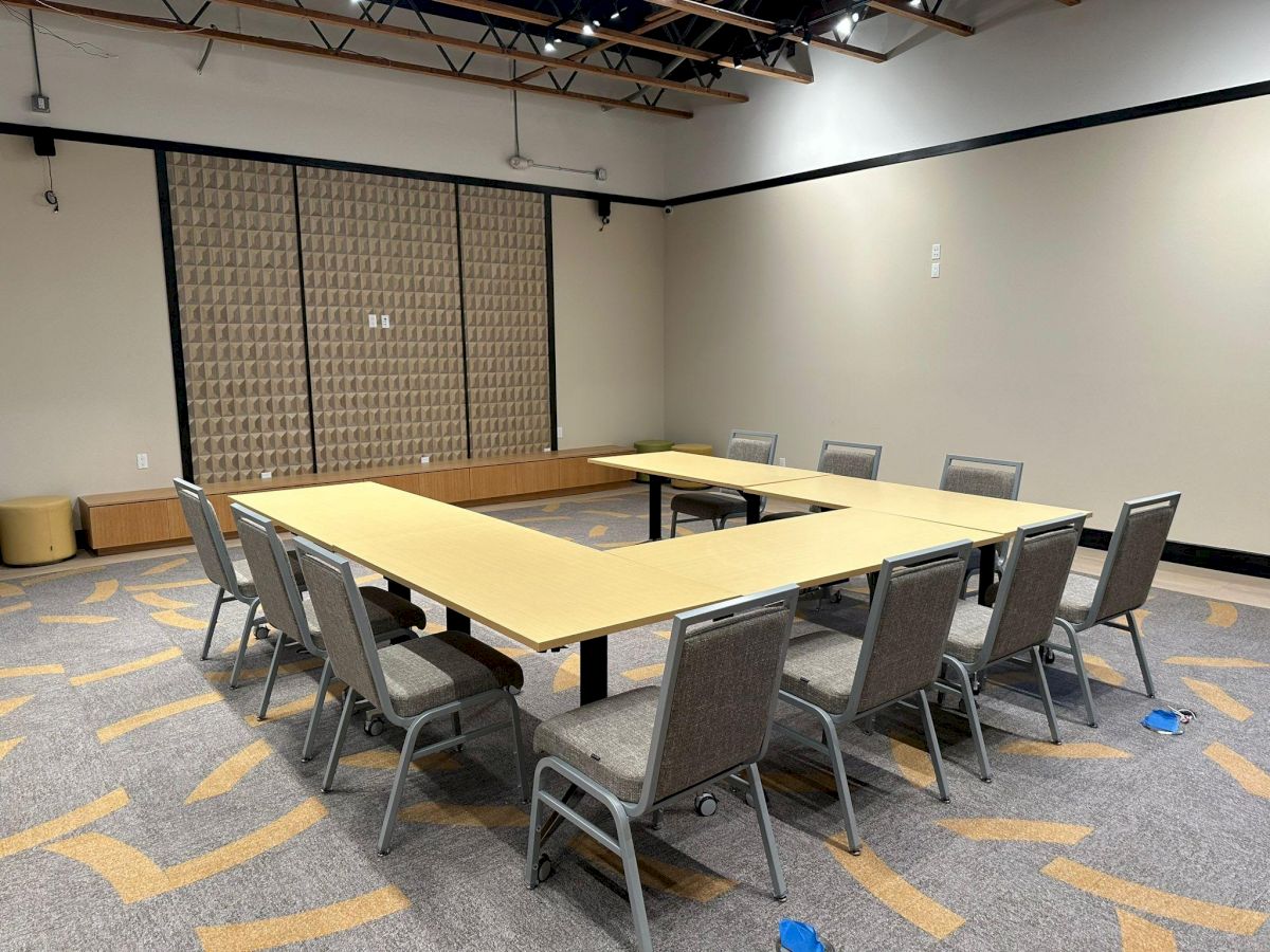 A conference room with a U-shaped table setup, surrounded by grey chairs on a patterned carpet floor. The walls are neutral, and the ceiling is exposed.