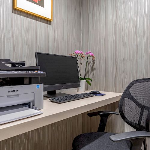 An office workspace with a printer, computer monitor, keyboard, mouse, office chair, and decorative plant on a table against a striped wall.