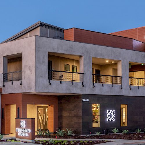 The image shows a modern building with signage indicating it's a hotel. The building has two floors, balconies, and landscaping in front.