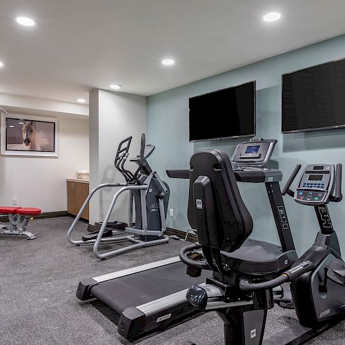 A small gym with exercise equipment, including a stationary bike, treadmill, elliptical machine, and weight machine, with two TVs mounted on the wall.