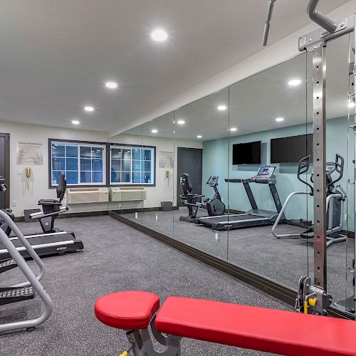 A modern gym with exercise equipment including treadmills, an elliptical machine, and a bench press. A large mirror spans one wall.