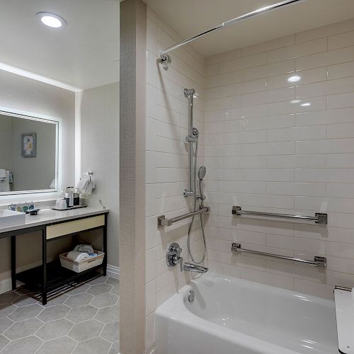 The image shows a modern bathroom with a sink and mirror on the left and a bathtub with grab bars and a shower on the right.