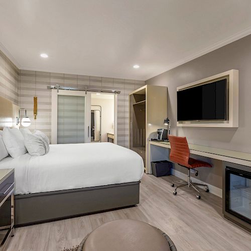 A modern hotel room with a large bed, desk with chair, TV, mini-fridge, closet, and stylish decor with ample lighting.