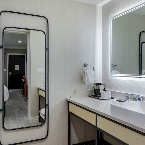 The image shows a modern bathroom with a large mirror, a sink, a towel rack, and a coffee maker next to the sink on the counter.