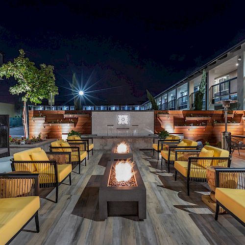 An outdoor seating area with yellow cushioned chairs surrounding fire pits, flanked by modern buildings at night.