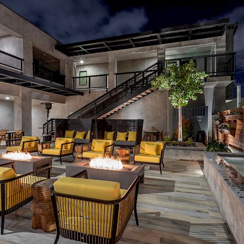 A modern outdoor patio area with yellow-cushioned lounge chairs, fire pits, tables, and decorative lighting next to a water feature.