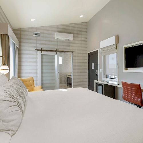 A modern hotel room with a large bed, a desk with a chair and TV, a small seating area, and a sliding door leading to the bathroom, well-lit naturally.