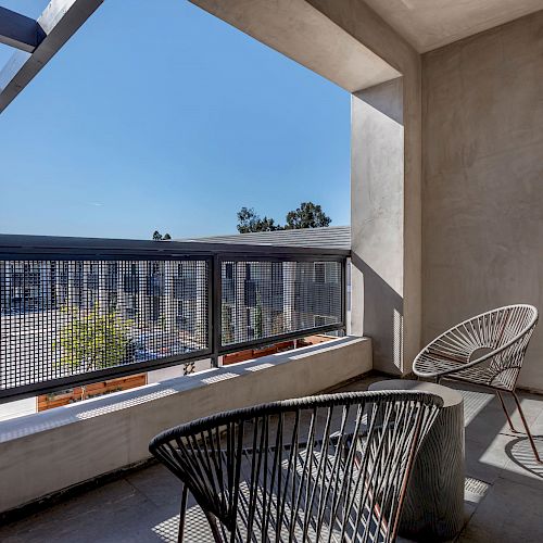 A modern balcony with two stylish chairs, a small table, and a view of the neighboring buildings under a clear blue sky.