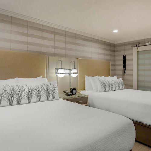 The image shows a modern hotel room with two neatly made double beds, decorative pillows, bedside lamps, and a sliding bathroom door, ending the sentence.