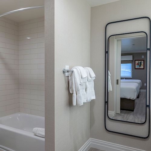 A bathroom with a bathtub and shower curtain on the left, towels on a rack, and a floor-length mirror reflecting a bed on the right wall.