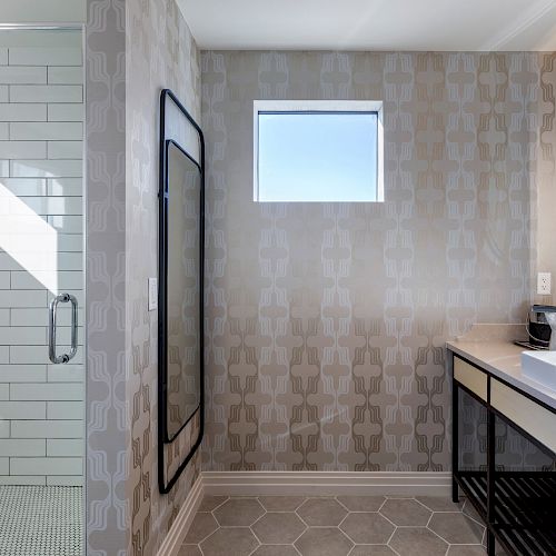 The image shows a modern bathroom with a glass-enclosed shower, a large mirror above a countertop with a sink, and a small window.