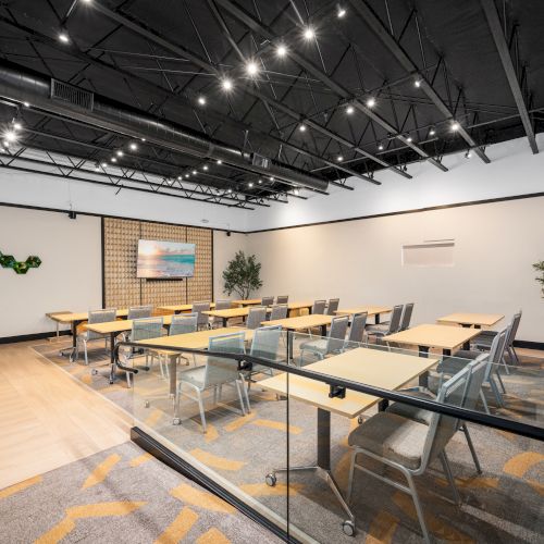 A modern conference room with tables, chairs, wall art, and plants, under a ceiling with exposed beams and lights.