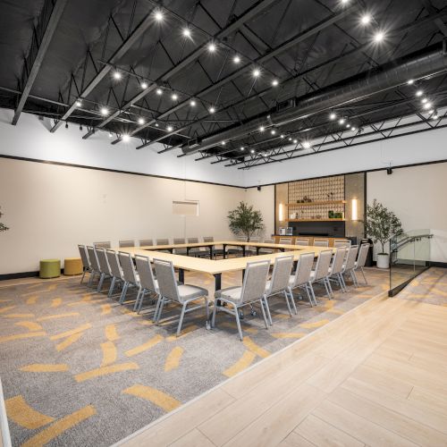 A modern conference room with a U-shaped table setup, chairs, plants, and a large TV screen, under stylish ceiling lights.