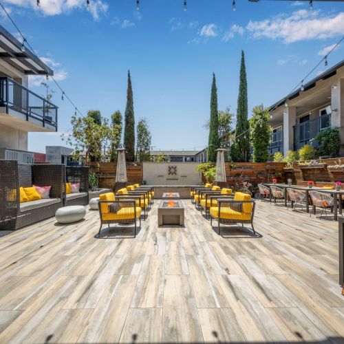 An outdoor seating area with cushioned chairs, tables, and string lights, flanked by greenery and modern architectural elements.
