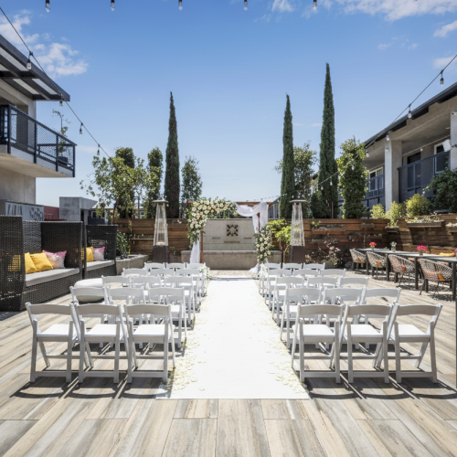 An outdoor wedding venue with white chairs, a central aisle, and string lights, set between modern buildings and greenery.