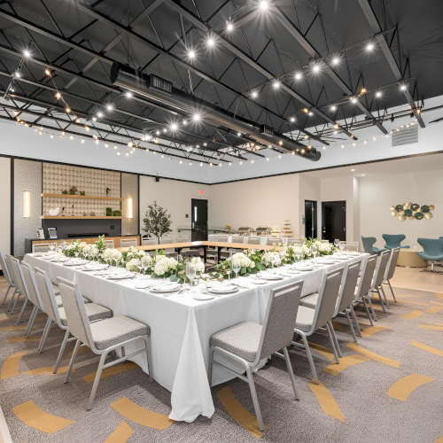 A well-lit event space features a U-shaped table setup with white tablecloths and floral centerpieces, modern decor, and overhead lighting.