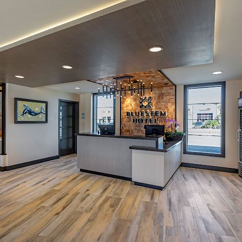 A modern hotel lobby with wood flooring, a reception desk, wall art, large windows, and a small convenience store section on the right.