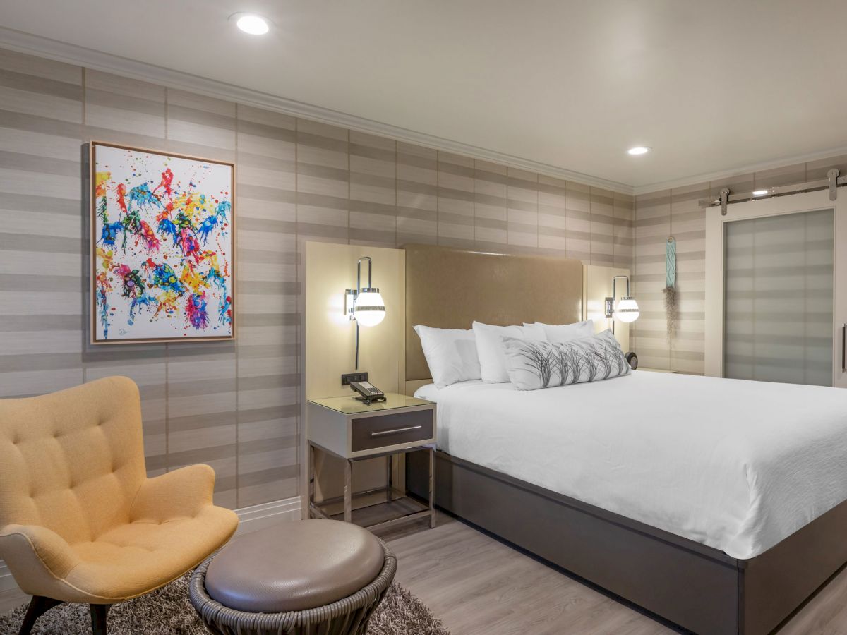 This image shows a neatly arranged hotel room with a double bed, a yellow chair, a round ottoman, and a colorful abstract painting on the wall.