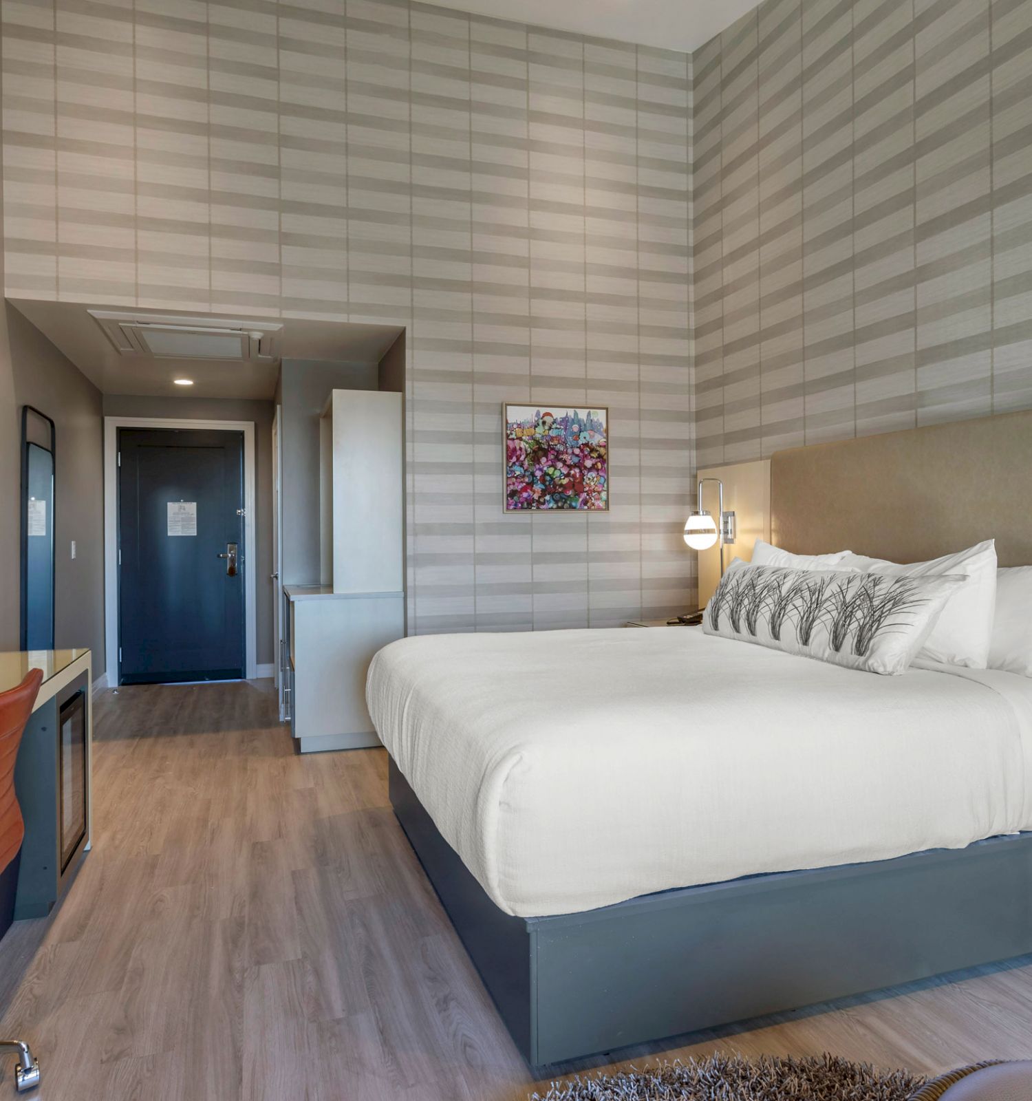 A modern hotel room features a large bed, desk with an orange chair, wall-mounted TV, lamps, and stylish decor in grey tones, creating a cozy atmosphere.