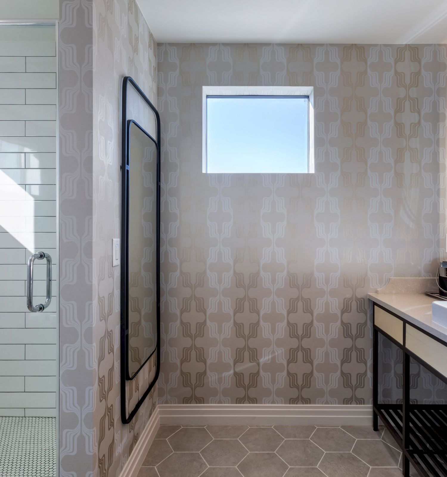 A modern bathroom with a glass-enclosed shower, patterned walls, a single window, a spacious vanity, and a large mirror above the sink.