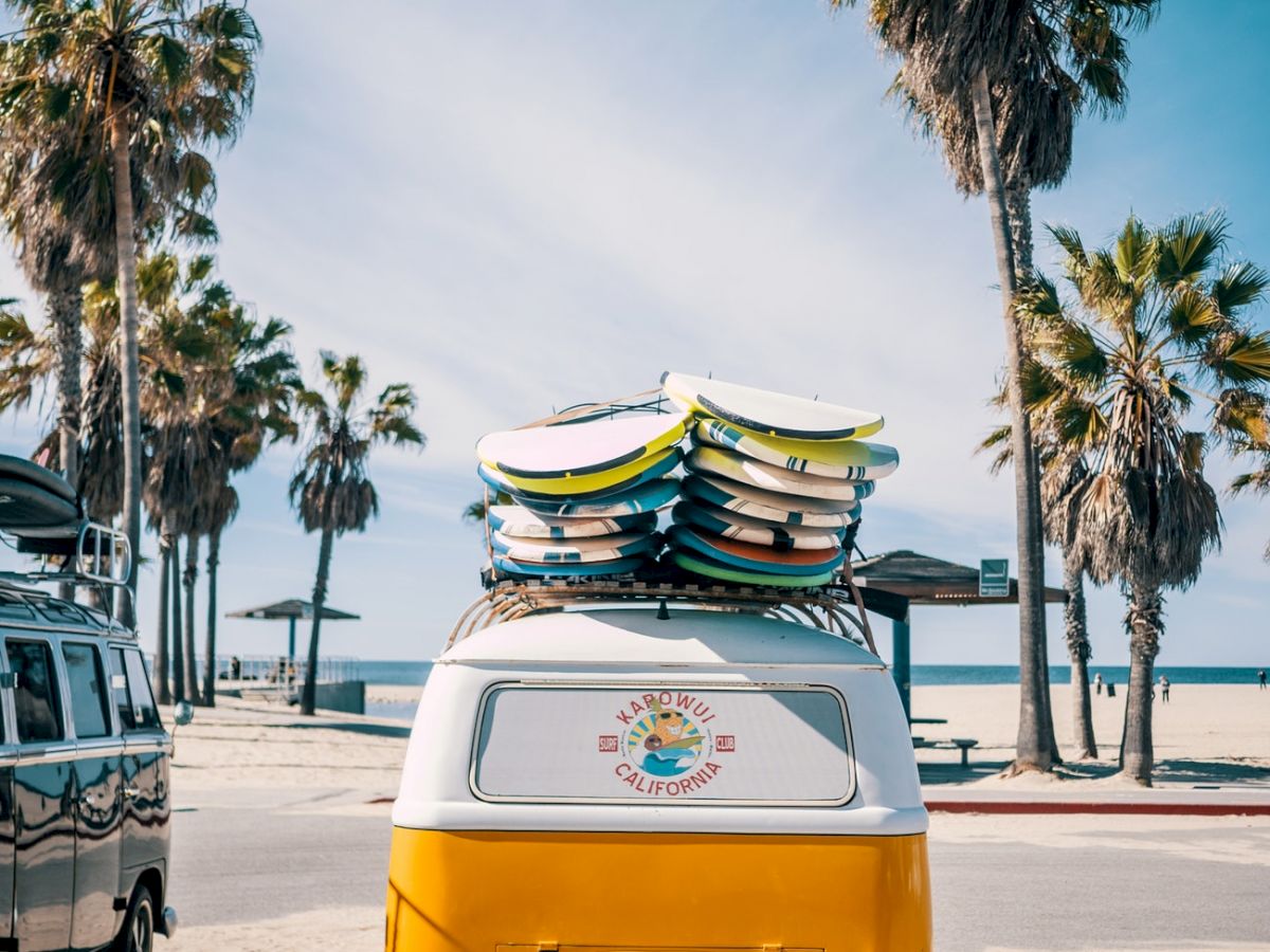 A yellow and white van with surfboards on top is parked near palm trees at a beach. The scene is sunny and captures a laid-back coastal vibe.