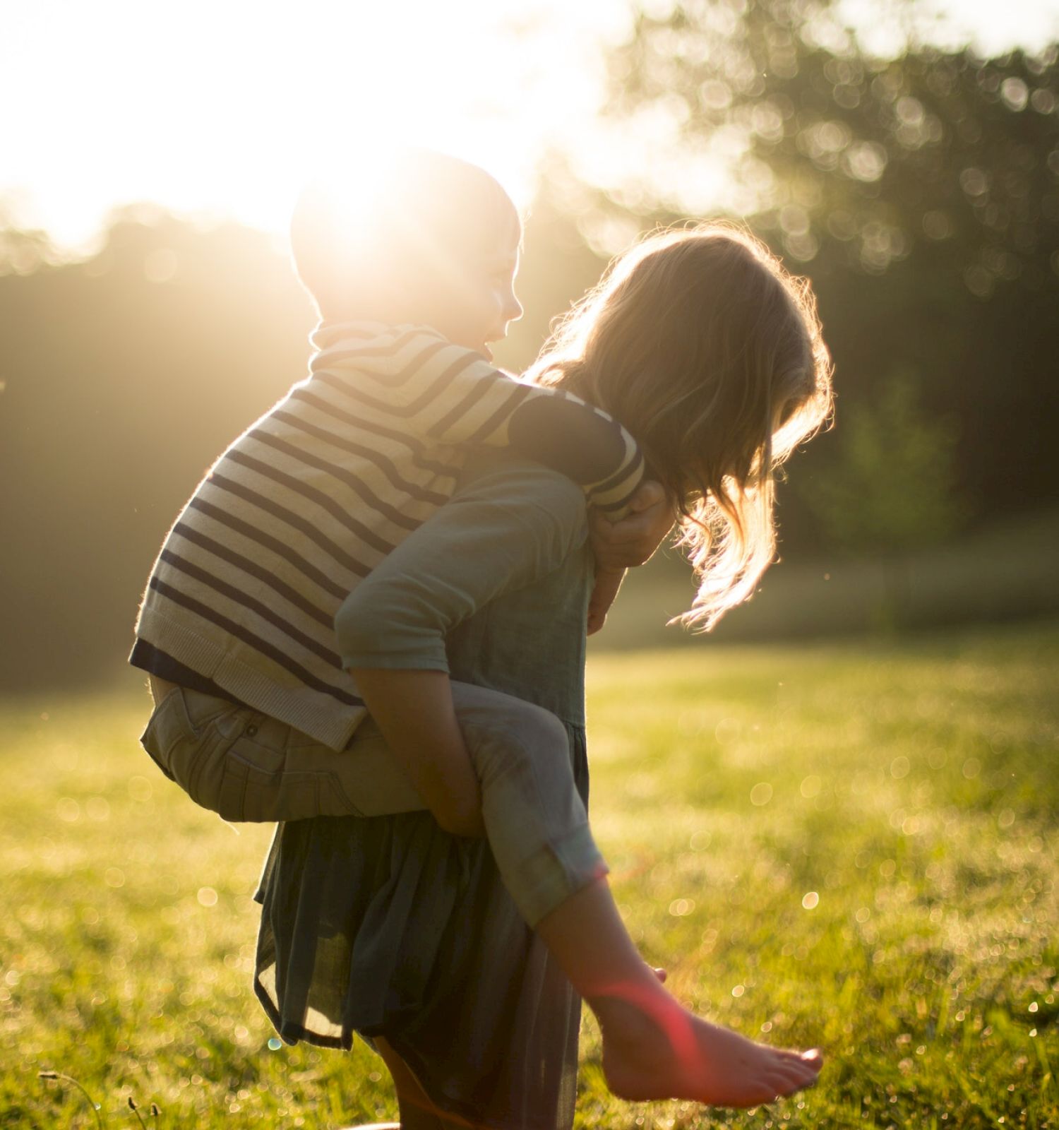 A child is giving another child a piggyback ride in a sunny field during sunset, enjoying a playful moment outdoors.