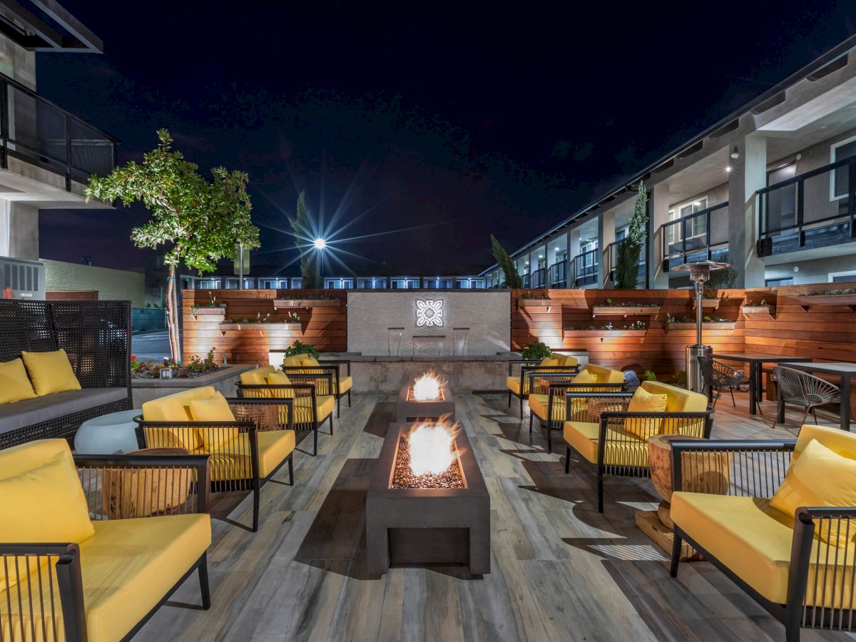 An outdoor lounge area at night with chairs around fire pits, modern furniture, yellow cushions, and surrounding buildings and greenery, seen lit up.