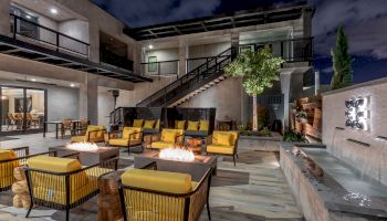 An outdoor patio at night with fire pits, yellow-cushioned chairs, a water feature, and modern architecture, featuring a staircase and a second level.